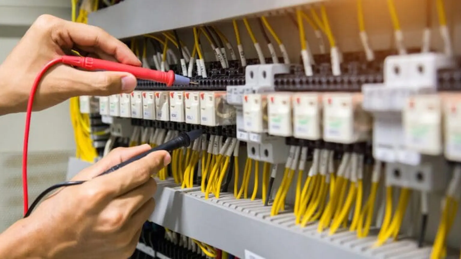 A technician performing 24-hour electrical repair by using a multimeter to test wiring and components in a complex industrial circuit breaker panel.