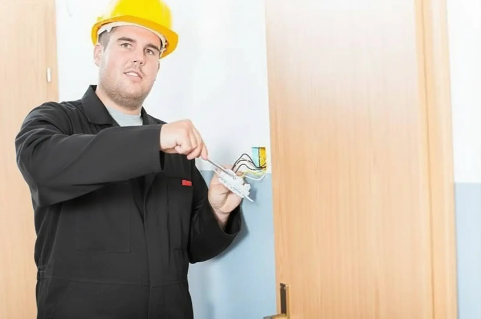 Electrical panel: An electrician wearing a yellow hard hat and black coveralls uses a screwdriver to wire a light switch into a wall outlet during a home renovation.