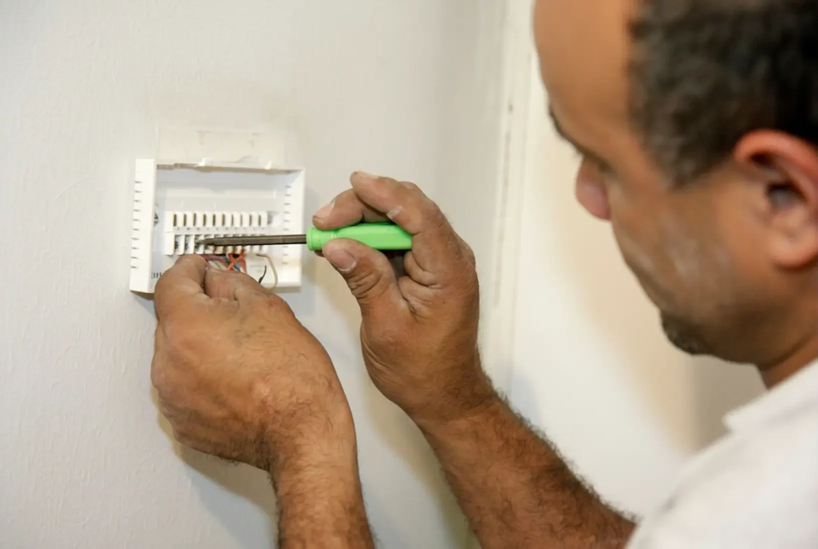 Close-up of an electrician's hands providing fast repairs, fixing a digital thermostat unit in a residential home.
