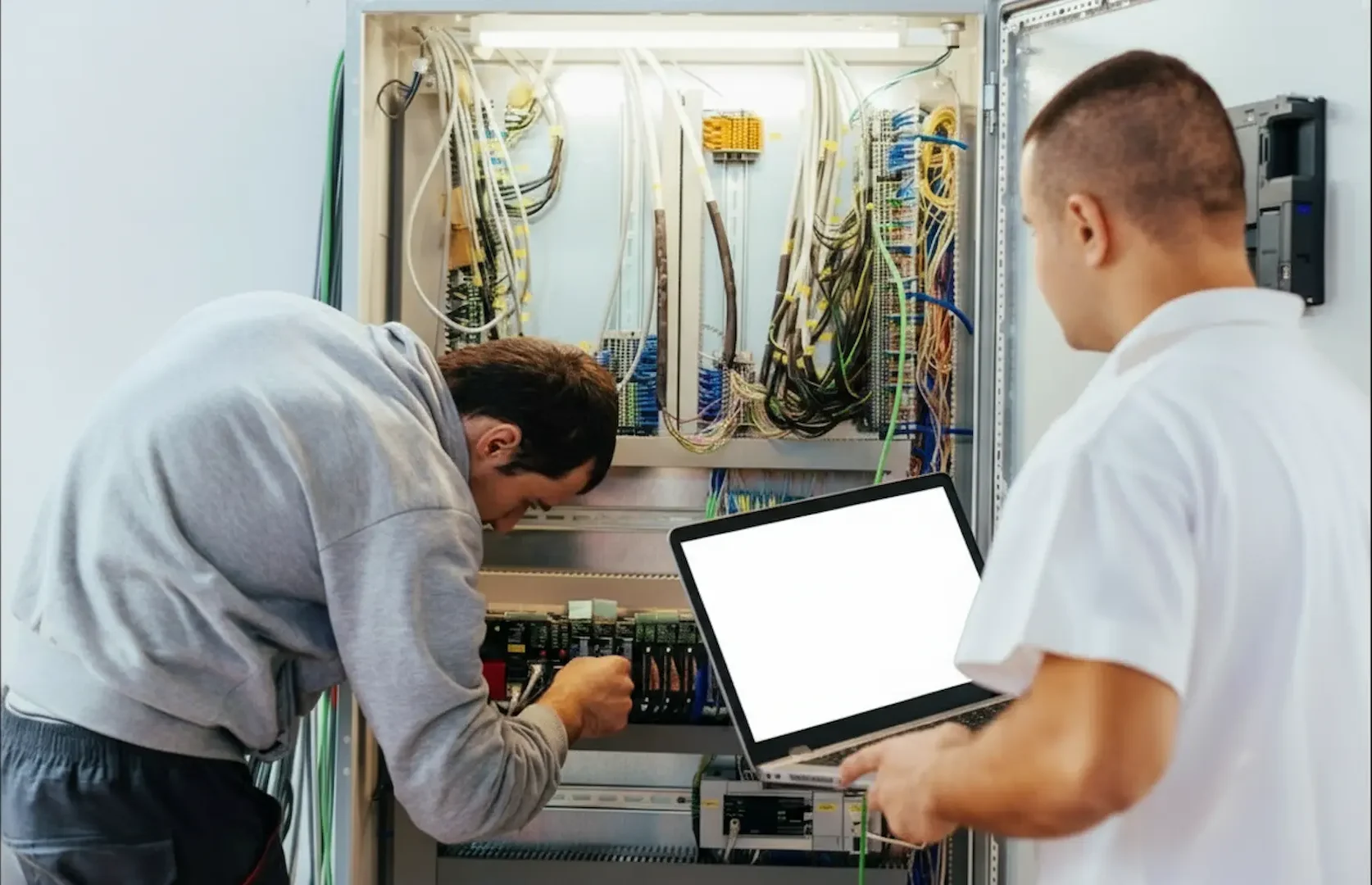 Two technicians working on an electrical panel upgrade, with one electrician adjusting internal wiring while the other monitors diagnostics on a laptop.