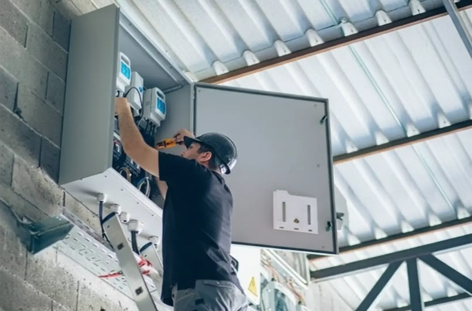 Immediate Electrical Assistance:A technician on a ladder works inside a large, wall-mounted industrial electrical panel located in a warehouse facility with corrugated roofing.