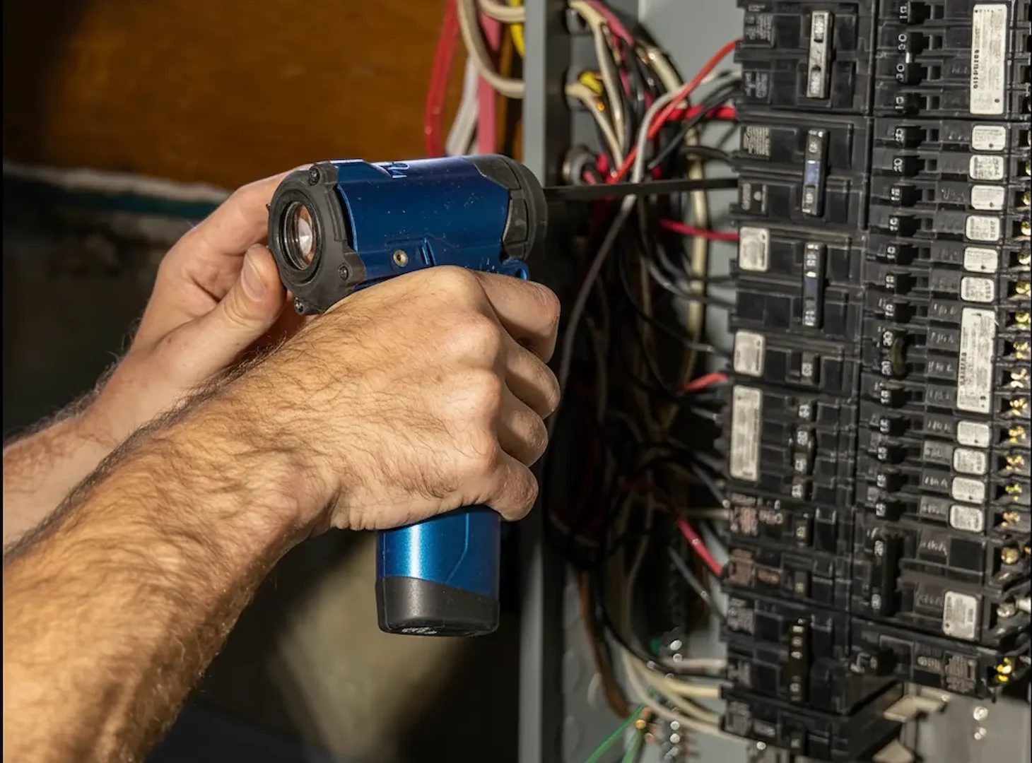 Cost of panel upgrades: An electrician in red safety gloves installs a new circuit breaker in a residential fuse box using needle-nose pliers, with a digital multimeter and tool pouch in the foreground.
