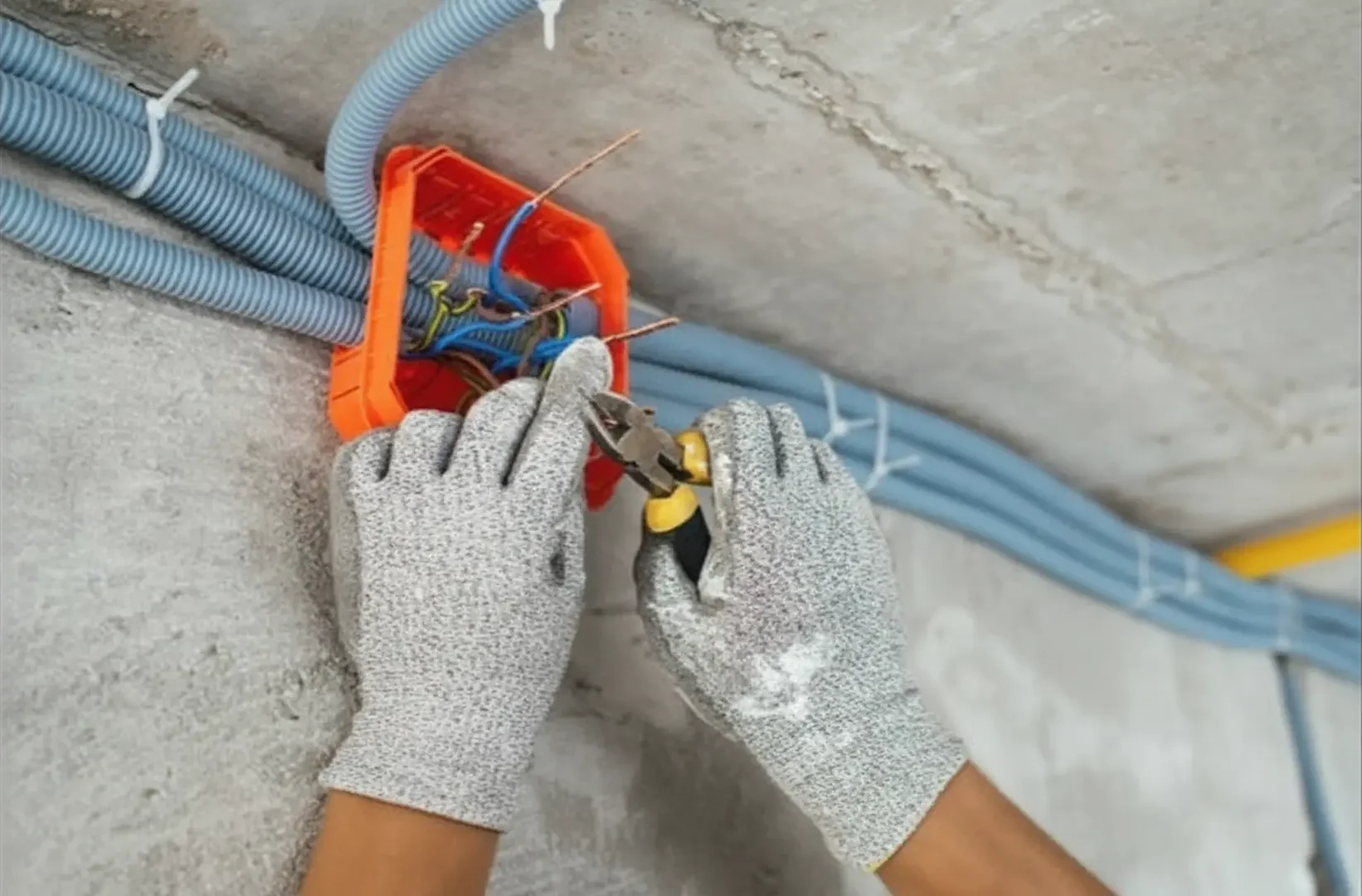 Cost of Panel upgrade: Electrical Panel: Close-up of an electrician's hands in protective gloves using wire cutters to strip copper wires inside an orange junction box with blue corrugated conduits on a concrete wall.