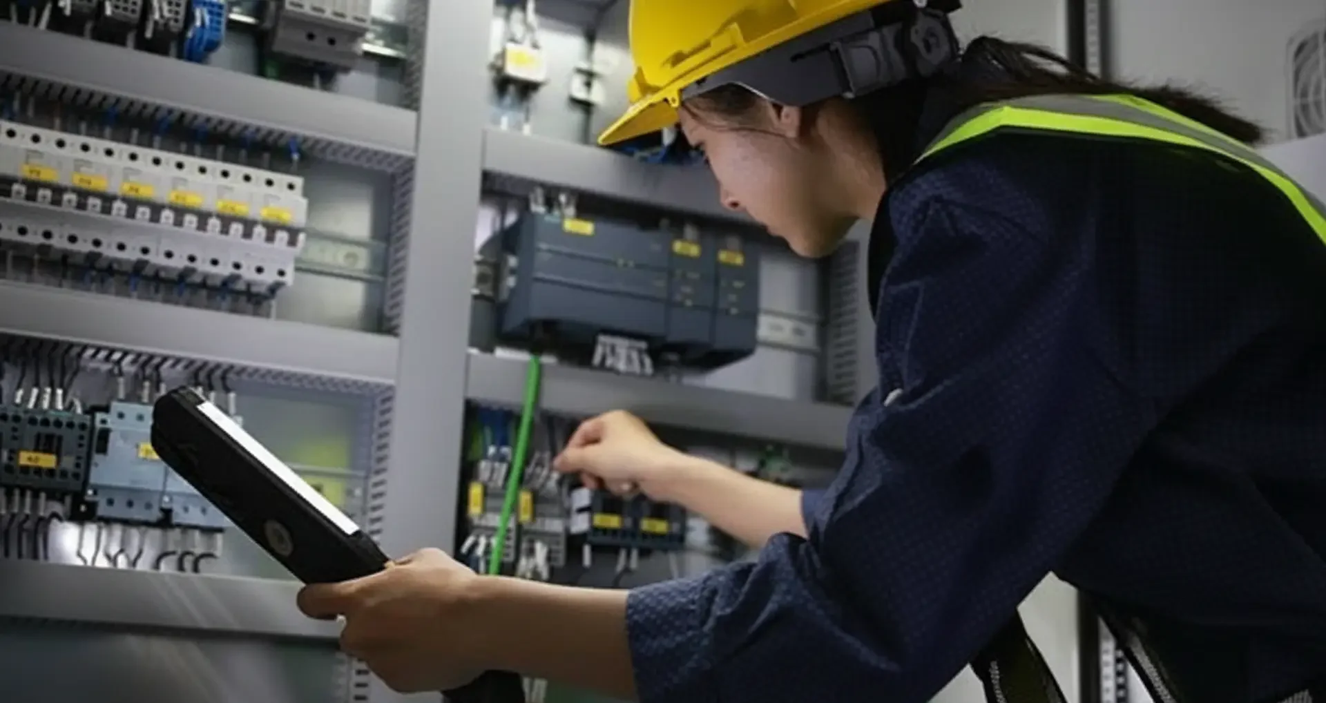 Emergency Electrical Service: A female technician in a yellow hard hat and safety vest uses a handheld diagnostic scanner to inspect intricate wiring inside a large-scale electrical panel.