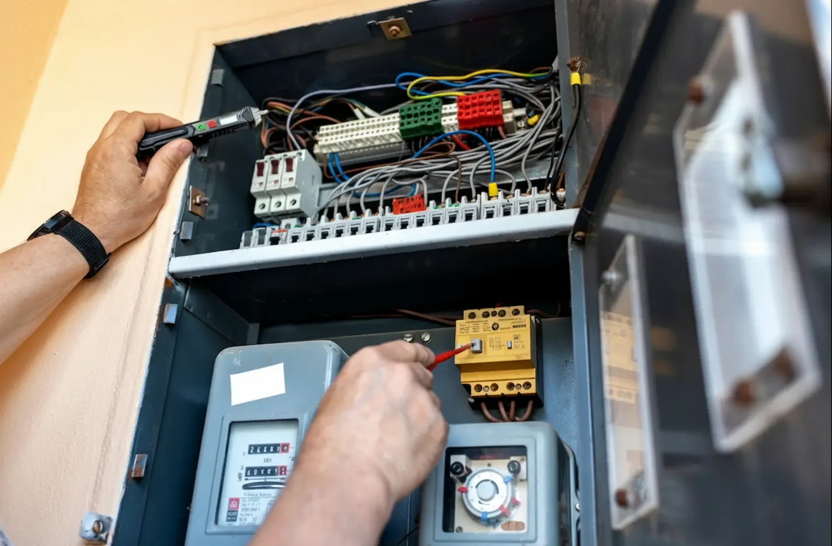 Electric Repair Cost: A close-up view of a technician's hands performing maintenance on an electrical service panel. One hand uses a digital voltage tester on upper terminal blocks, while the other hand uses a red insulated tool to adjust a yellow component near an analog utility meter.