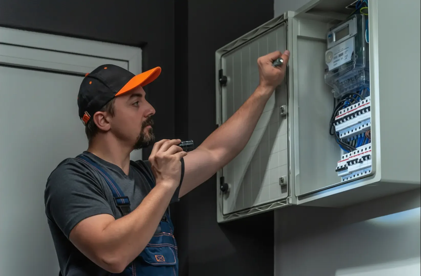 Electrician using a flashlight to inspect a digital meter and breakers in an open wall box.