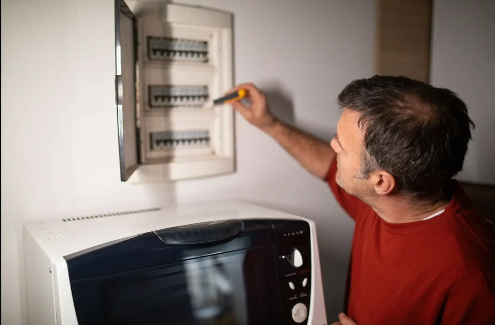 Electrical Emergency: A man in a red sweater using a yellow handheld tool to inspect or reset a white recessed electrical fuse box on a wall at night, located above a microwave oven.