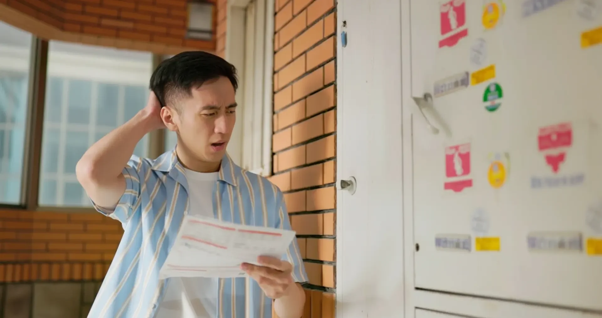 Booking an electrical repair: A concerned man in a striped blue shirt standing outdoors by a brick wall, scratching his head while looking at a paper utility bill in front of a white door with stickers.