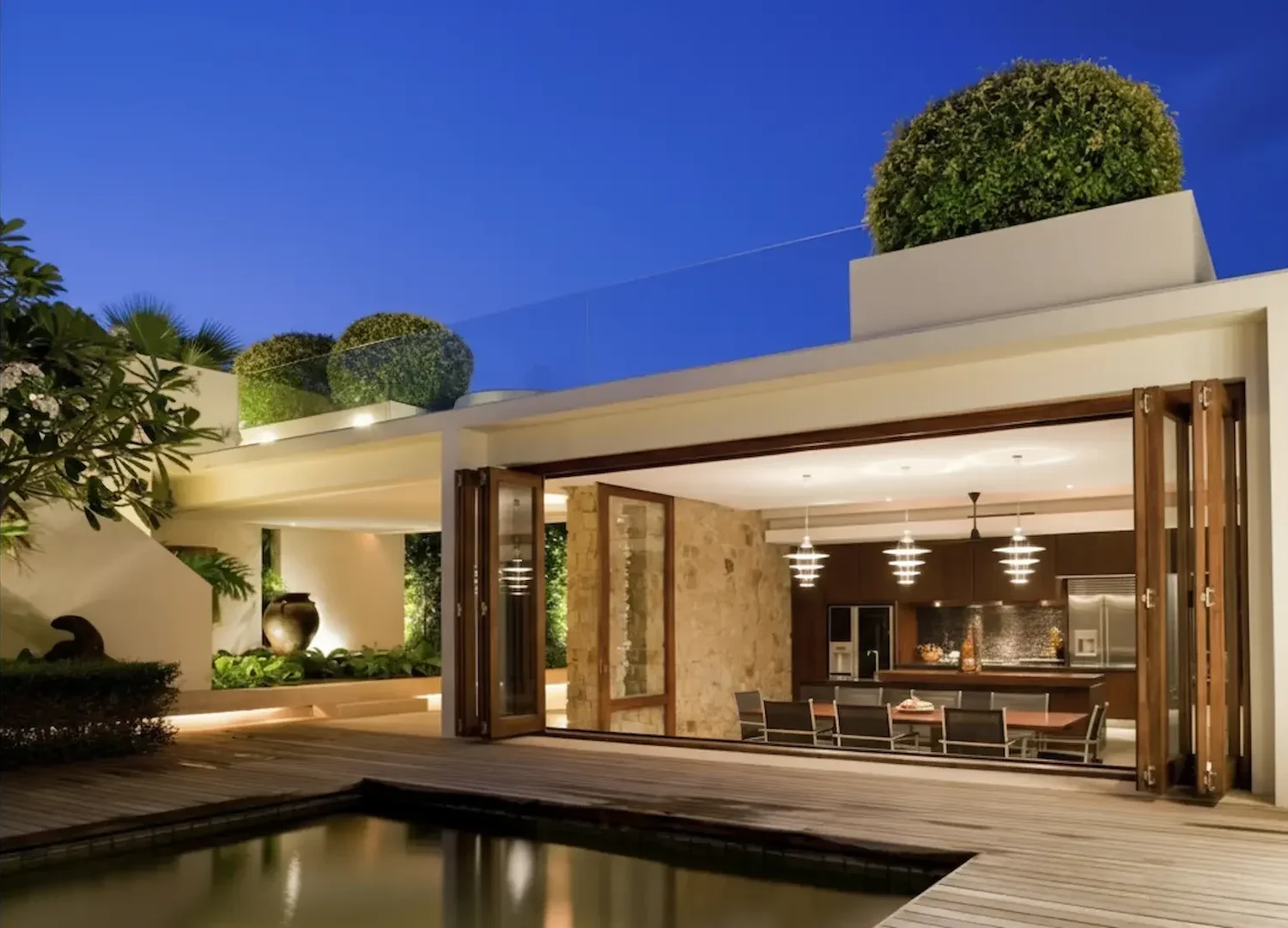 Outdoor Lighting design: A luxury poolside patio area at night with sliding glass doors, featuring hanging pendant lights in the kitchen and warm recessed deck lighting.