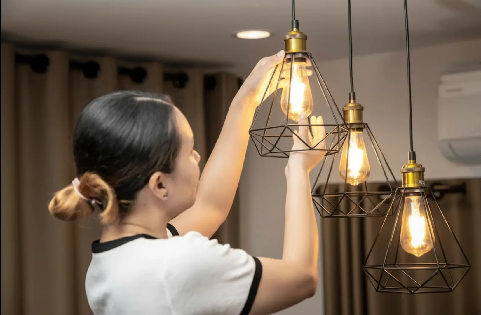 Local labor rates: A woman installing an Edison-style LED bulb into a black geometric wire-frame pendant light fixture.