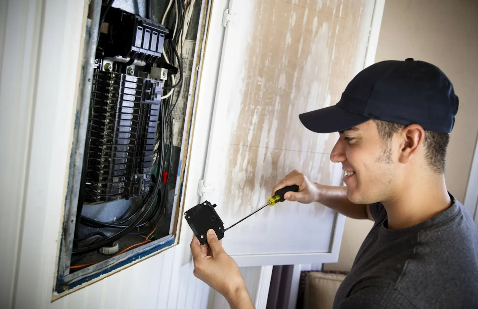 An electrician wearing a yellow hard hat, ear protection, and safety glasses uses pliers to manage wiring inside a wall-mounted panel with blue conduits.