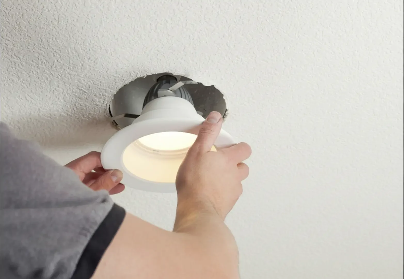 Emergency Electrician: Close-up of hands installing a white recessed LED ceiling light into a circular opening in a textured ceiling.