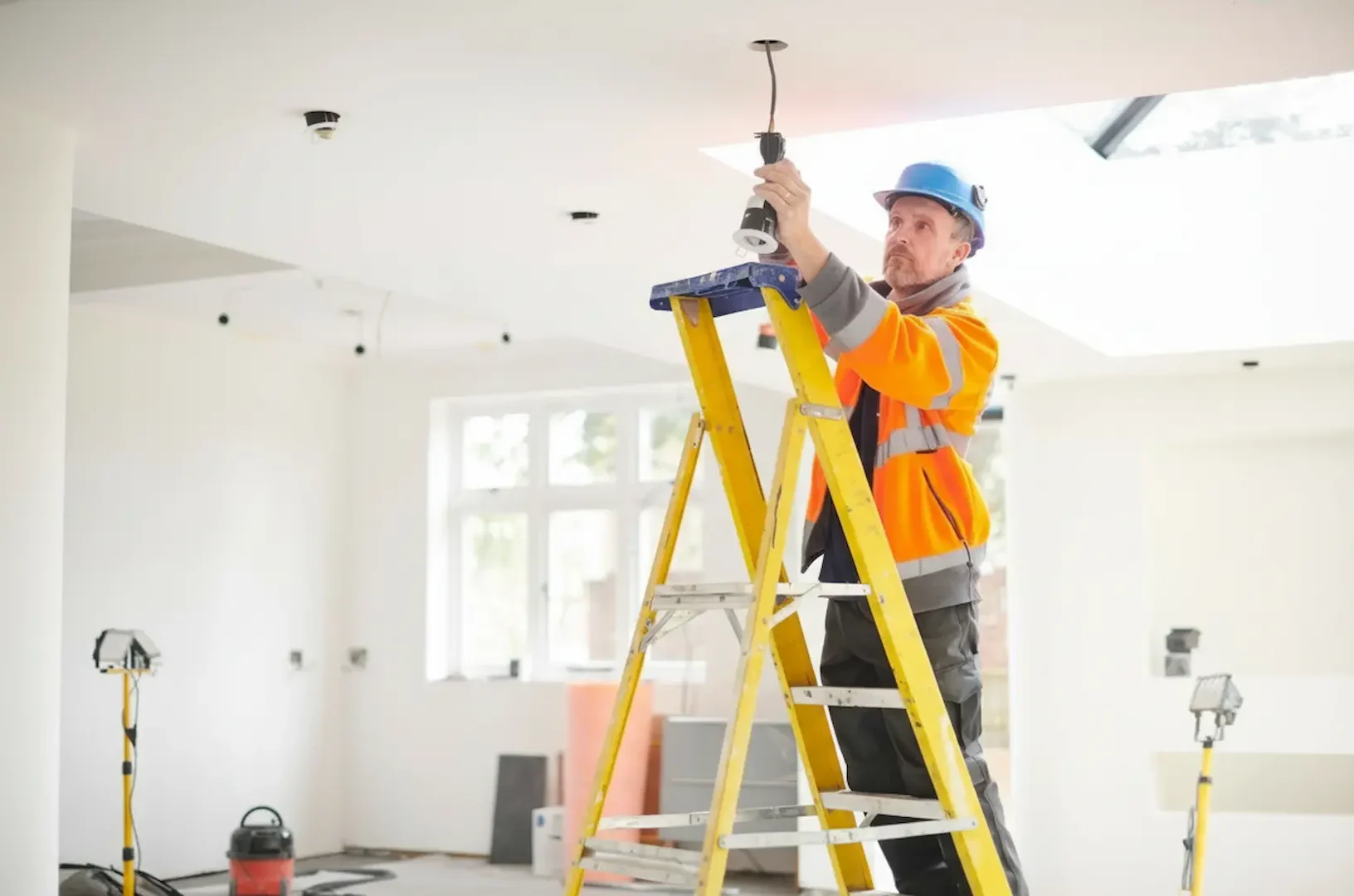 Electrical Service: Electrician on a safety ladder installing recessed LED lighting fixtures in a high-ceiling residential living space.