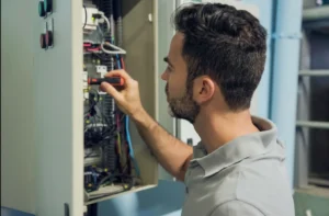 Electrician in Stony Brook, NY: A Guide to Repairs, Older Homes & Electrical Upgrades 5 Electric repair cost: A side view of a male electrician with a beard, wearing a grey polo shirt, using a precision screwdriver to carefully adjust or repair internal wiring components inside an open industrial electrical control cabinet filled with complex circuitry, switches, and multicolored wires.