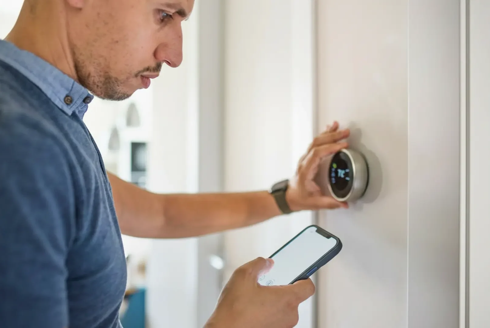 Booking an Electrical Repair: A man in a blue shirt standing in a hallway, holding a smartphone while adjusting a circular silver smart thermostat mounted on the wall.