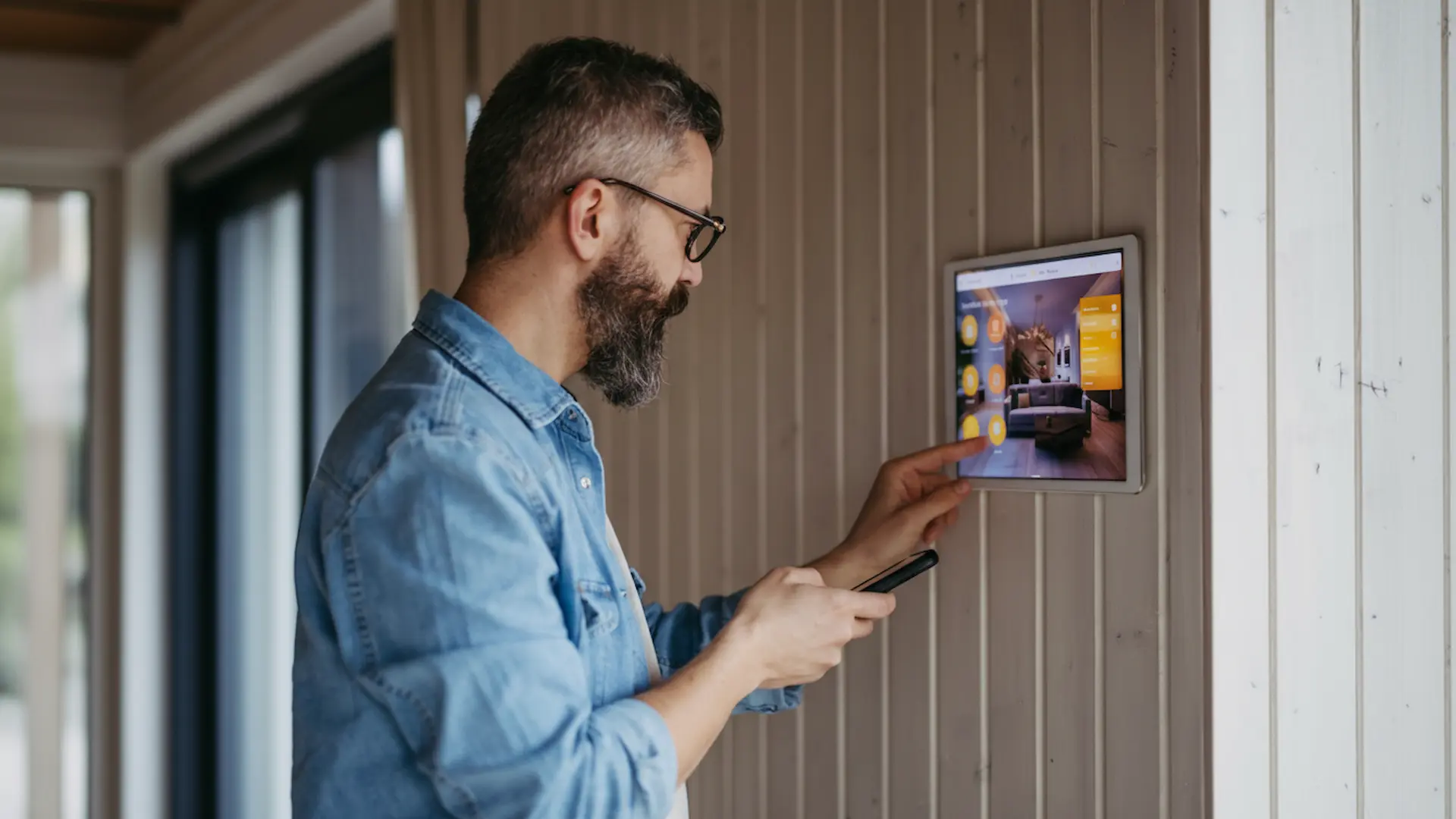 Emergency electrical Service: A man with a beard and glasses using a smartphone while interacting with a large smart home control panel mounted on a wood-paneled wall.